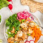 A plate with cooked salmon bites, sliced radishes, pickled onions, shredded carrots, edamame, cilantro, lime wedge, and a sprinkle of sesame seeds, arranged on a white dish.