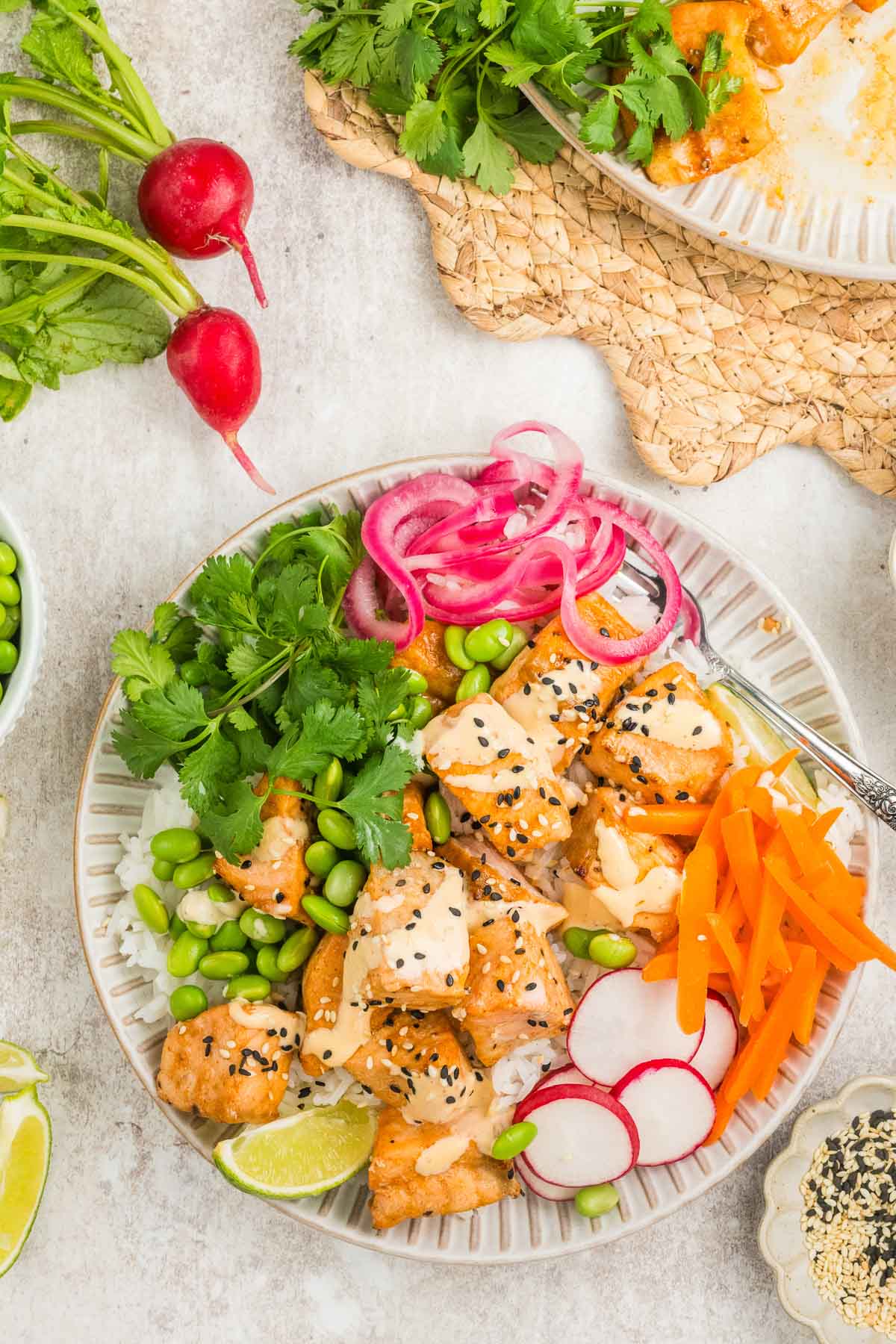 A plate with cooked salmon bites, sliced radishes, pickled onions, shredded carrots, edamame, cilantro, lime wedge, and a sprinkle of sesame seeds, arranged on a white dish.