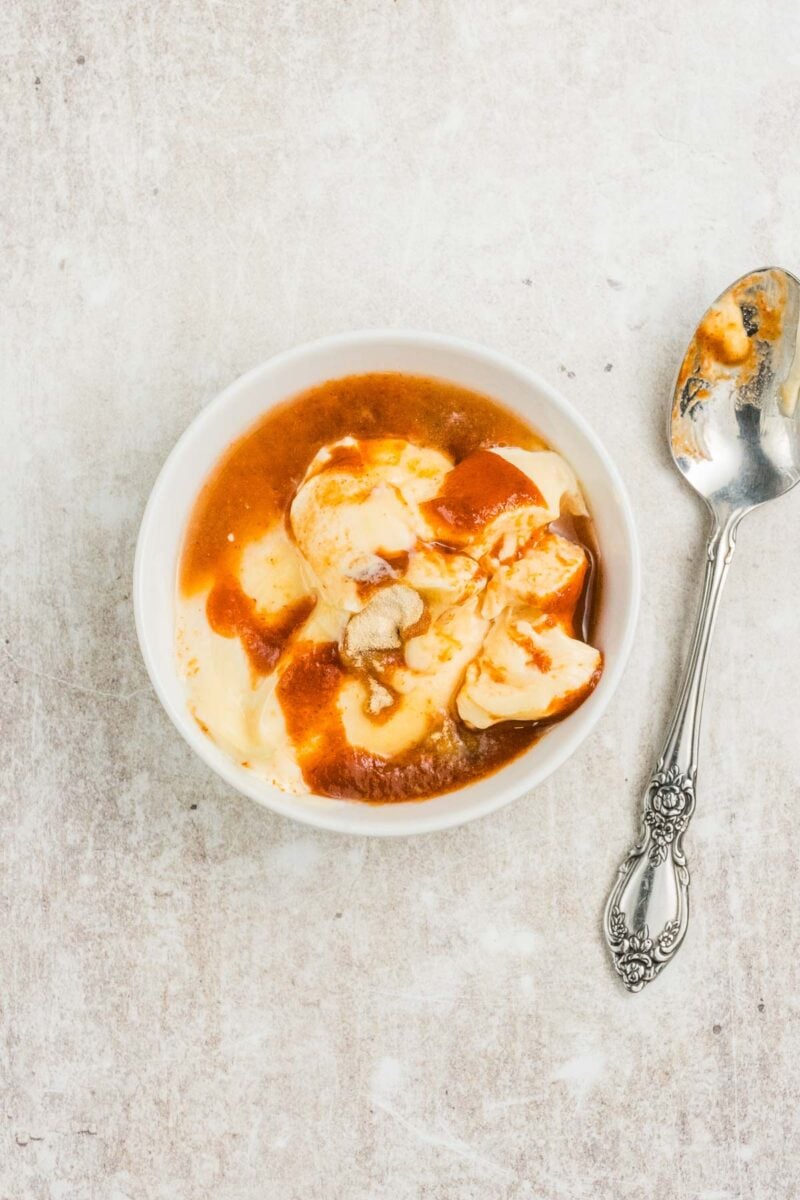 A white bowl of vanilla ice cream topped with caramel sauce sits next to a silver spoon on a light-colored surface.