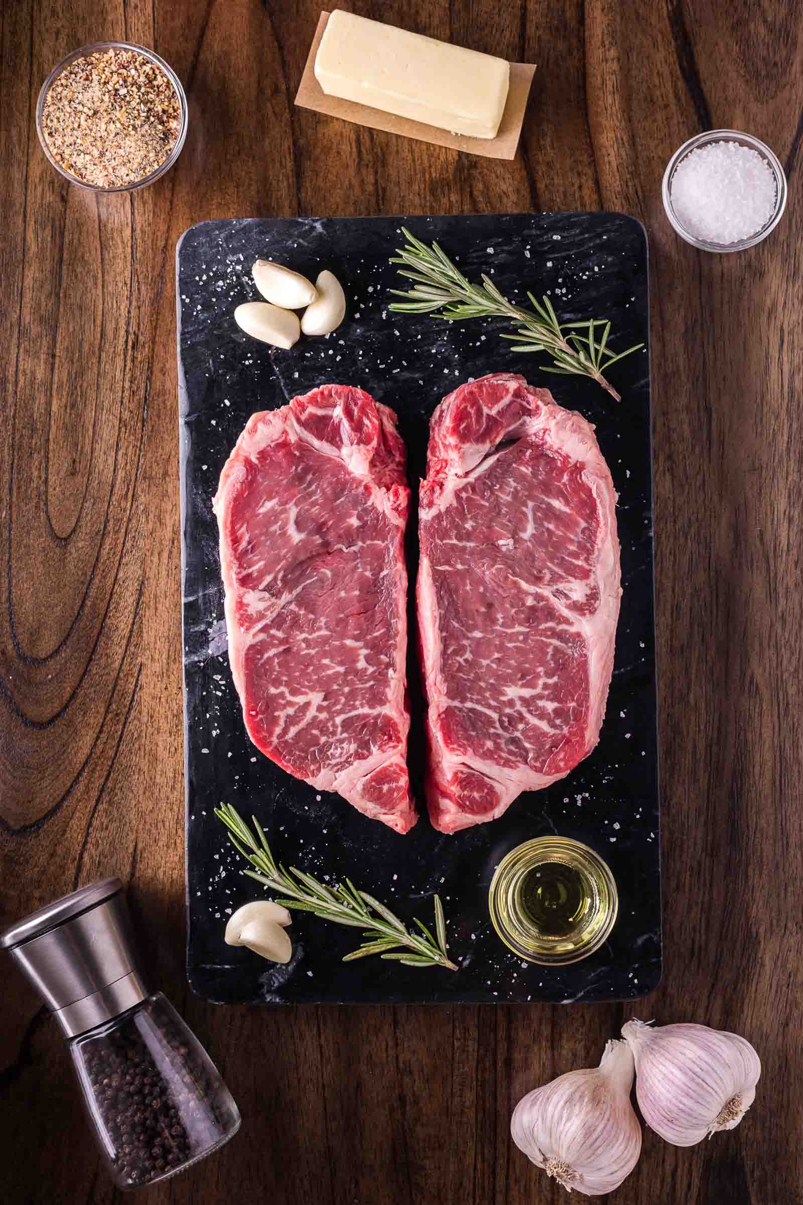 Two raw, marbled steaks on a black slate with rosemary, garlic, oil, butter, salt, pepper, and spices arranged around them on a wooden surface.