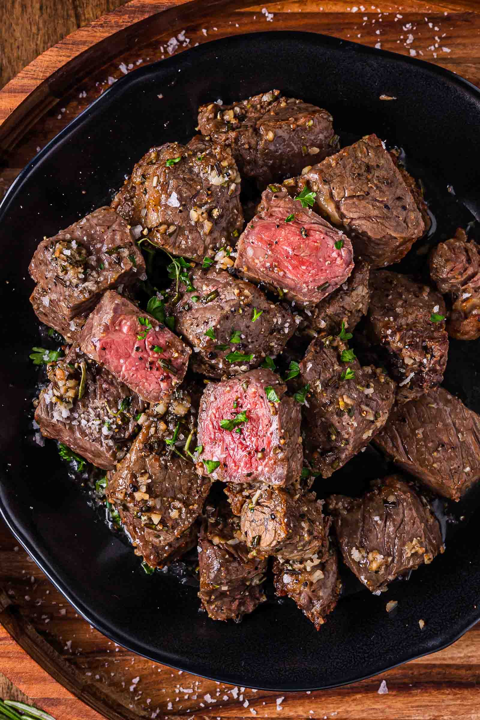 A black plate filled with garlic butter steak bites, some medium-rare, garnished with chopped herbs and coarse salt.