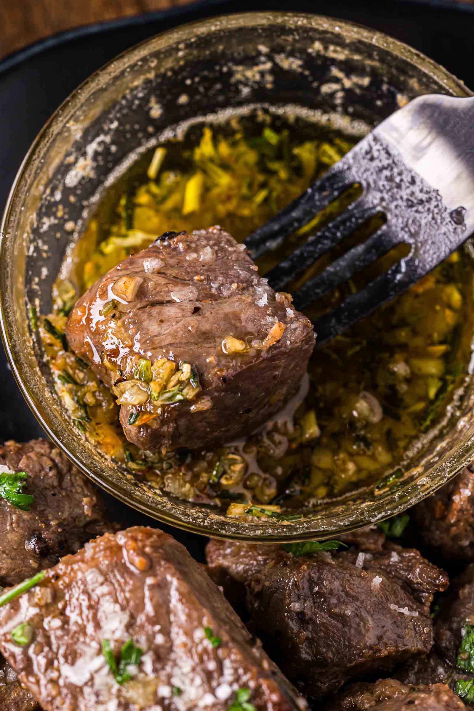 A fork holds a piece of cooked steak being dipped into a small glass bowl of garlic butter sauce, with more steak pieces on a plate below.