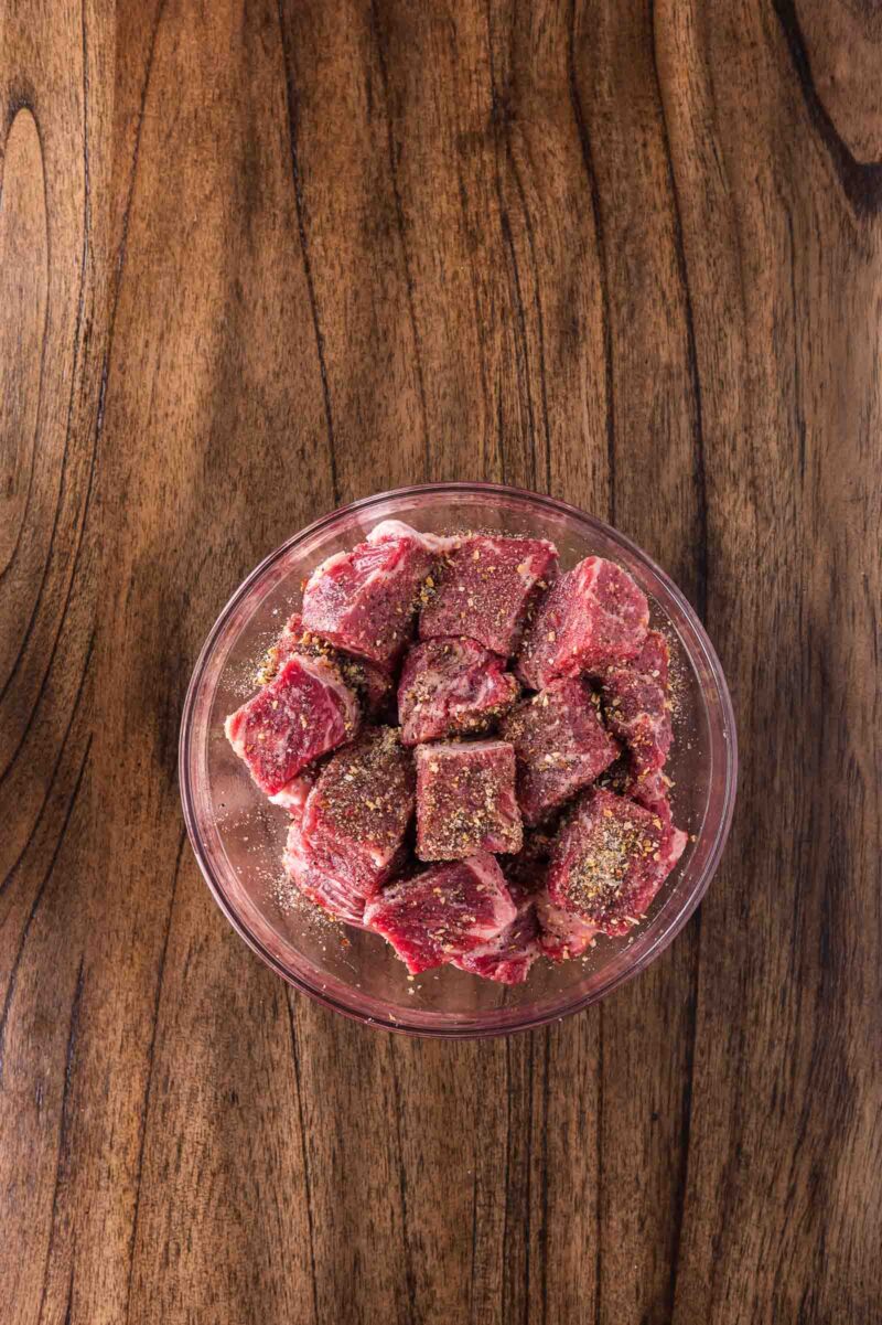 A glass bowl filled with seasoned raw beef cubes sits on a wooden surface.