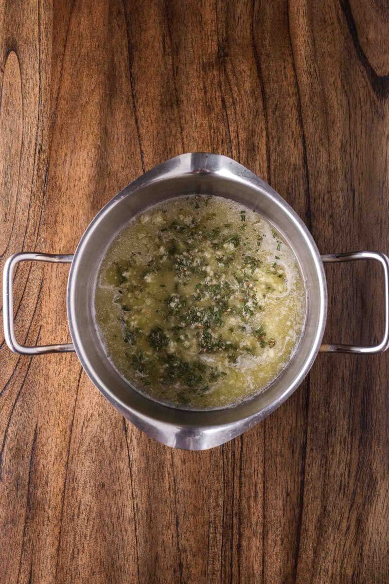 A stainless steel bowl with a melted butter and herb mixture sits on a wooden surface.