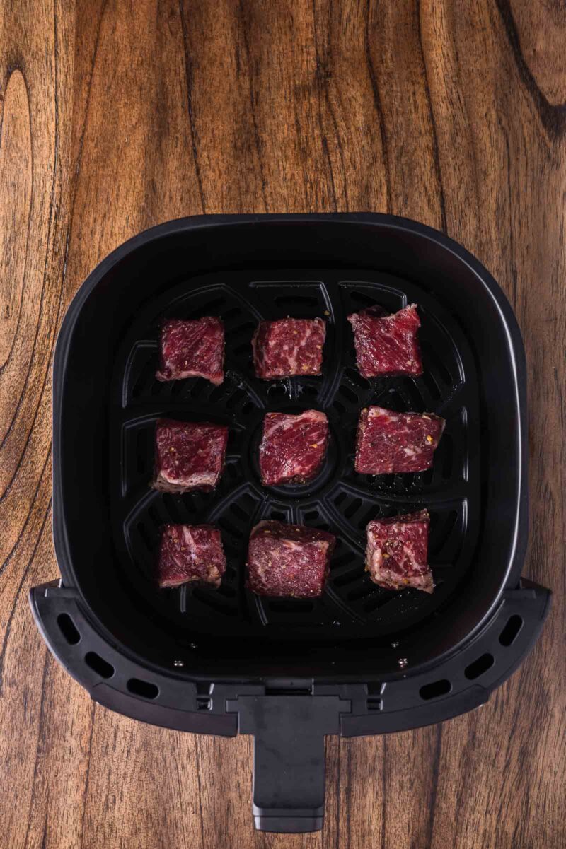 Nine pieces of raw steak are arranged inside the basket of an air fryer, which is placed on a wooden surface.