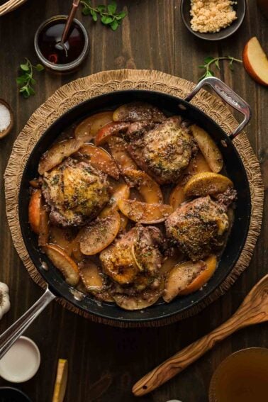 A skillet filled with cooked chicken thighs and sliced apples in a seasoned sauce, surrounded by small bowls of ingredients on a wooden table.