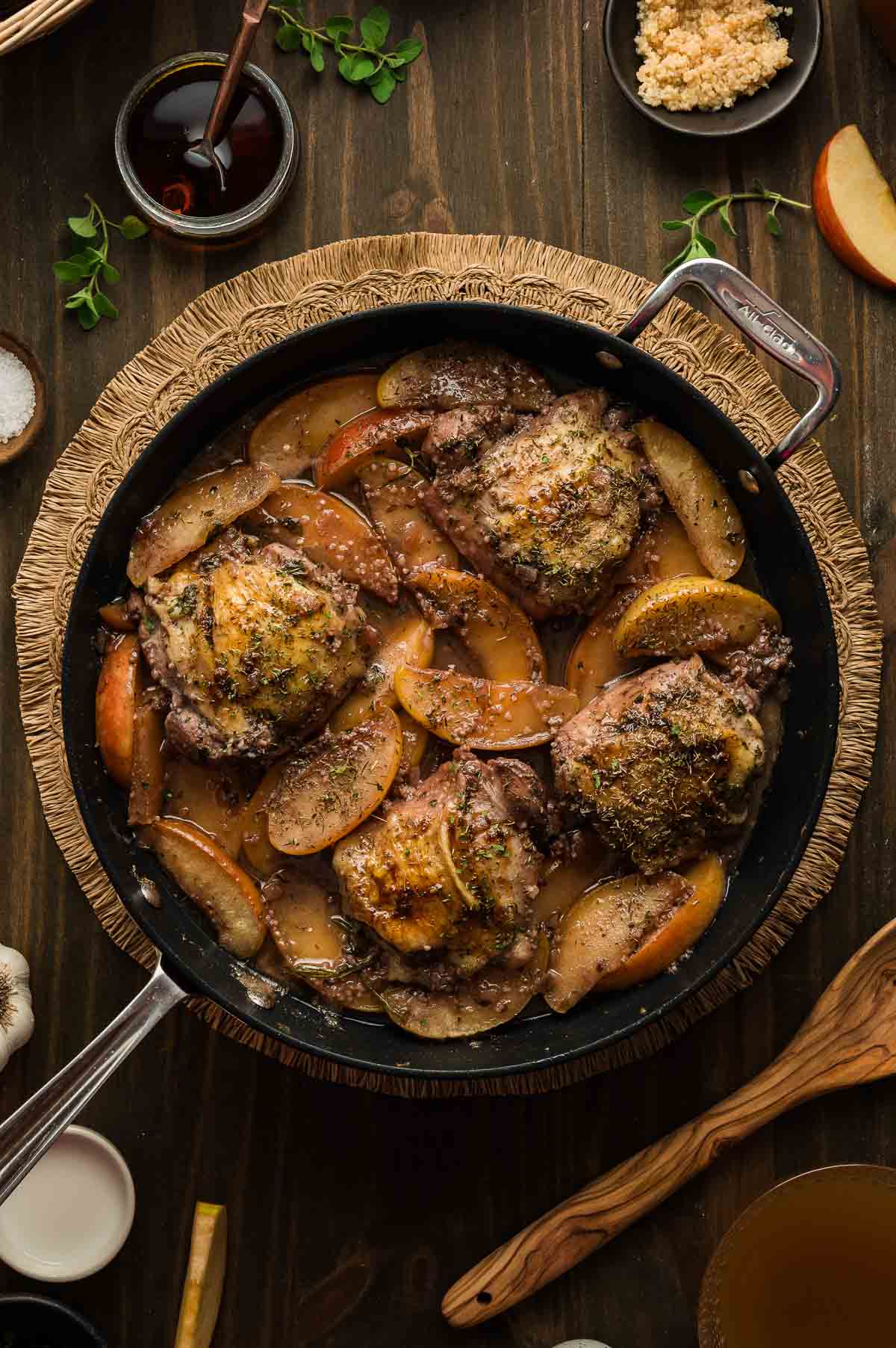 A skillet filled with cooked chicken thighs and sliced apples in a seasoned sauce, surrounded by small bowls of ingredients on a wooden table.