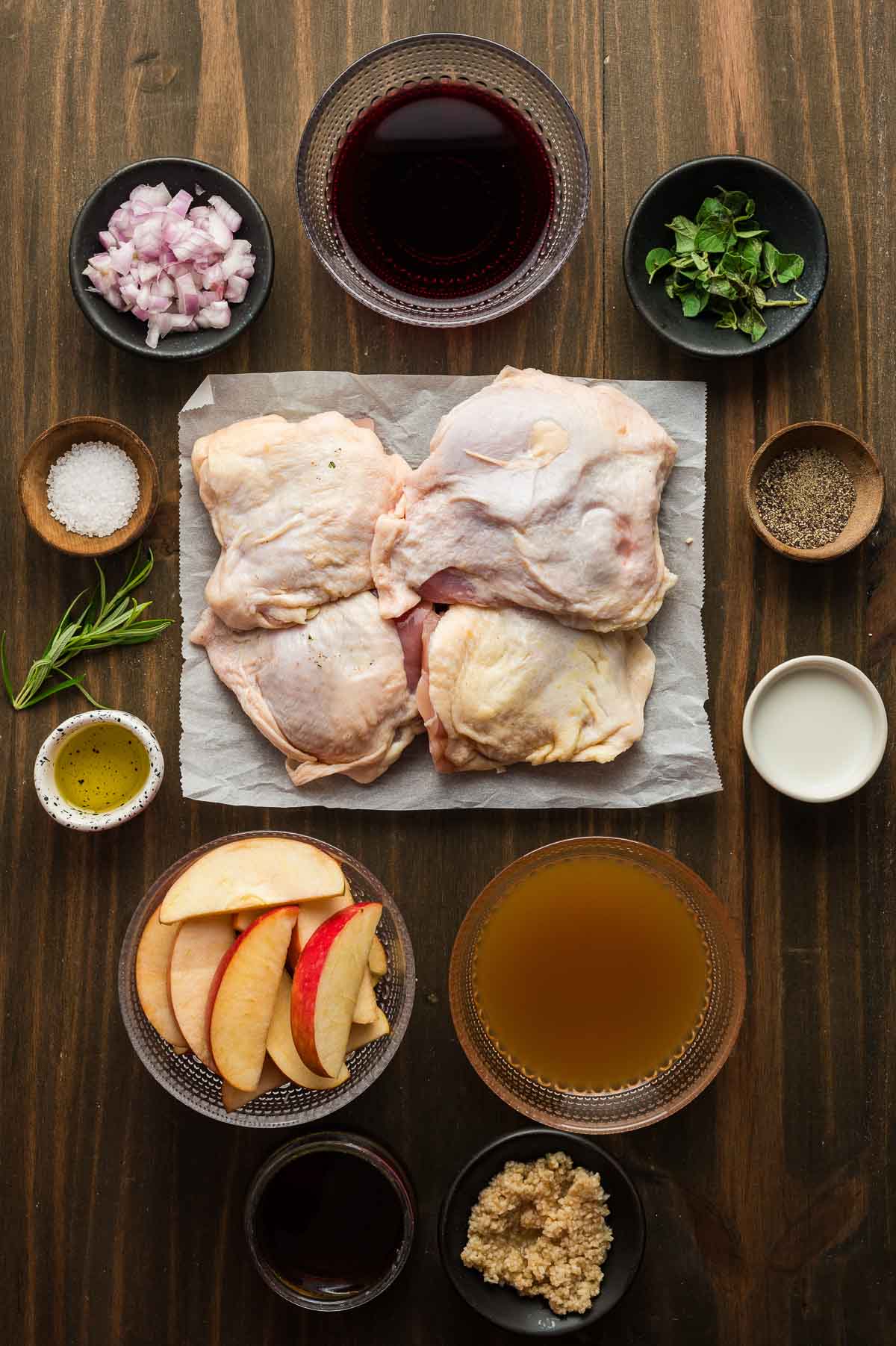 Raw chicken thighs on parchment surrounded by apple slices, chopped shallots, minced garlic, herbs, red wine, apple cider, olive oil, salt, and pepper on a wooden surface.