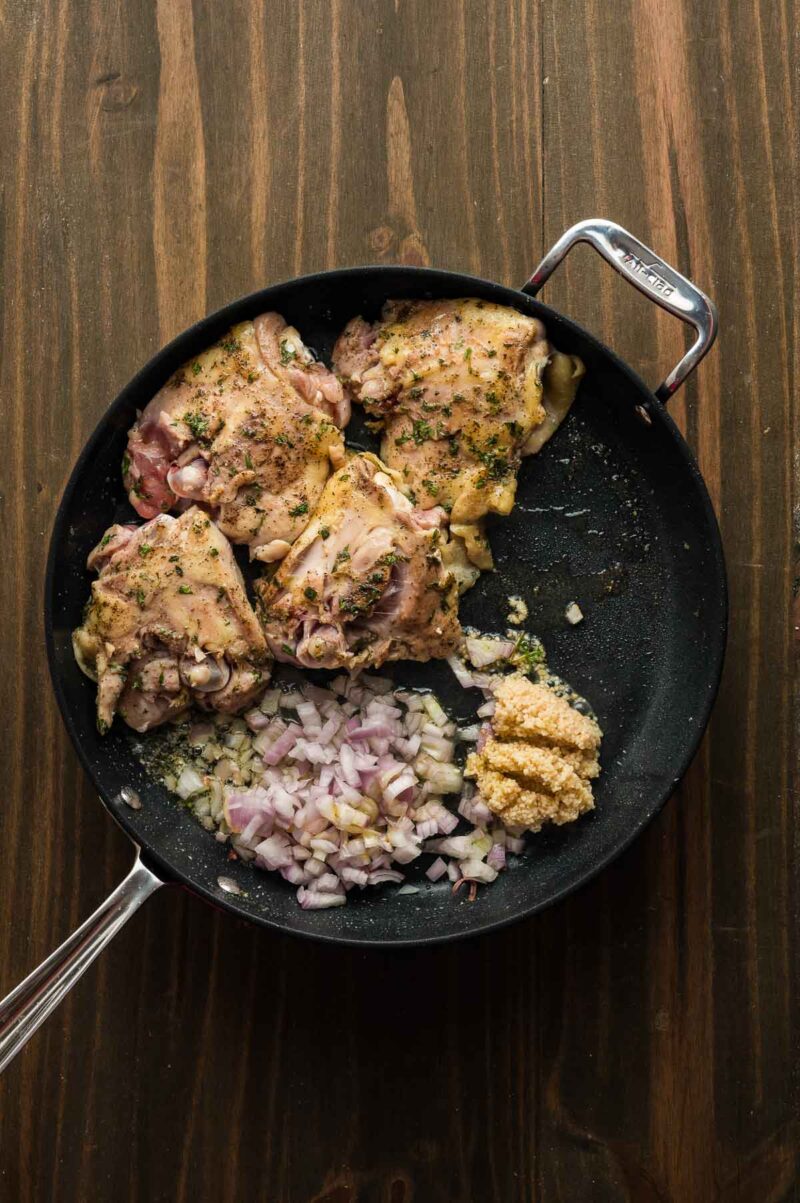 A skillet with seasoned raw chicken thighs, chopped onions, and minced garlic on a wooden surface.