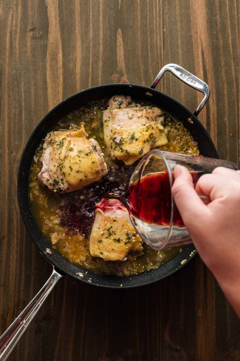A hand pours red wine into a skillet with seasoned chicken thighs simmering in a sauce on a wooden surface.
