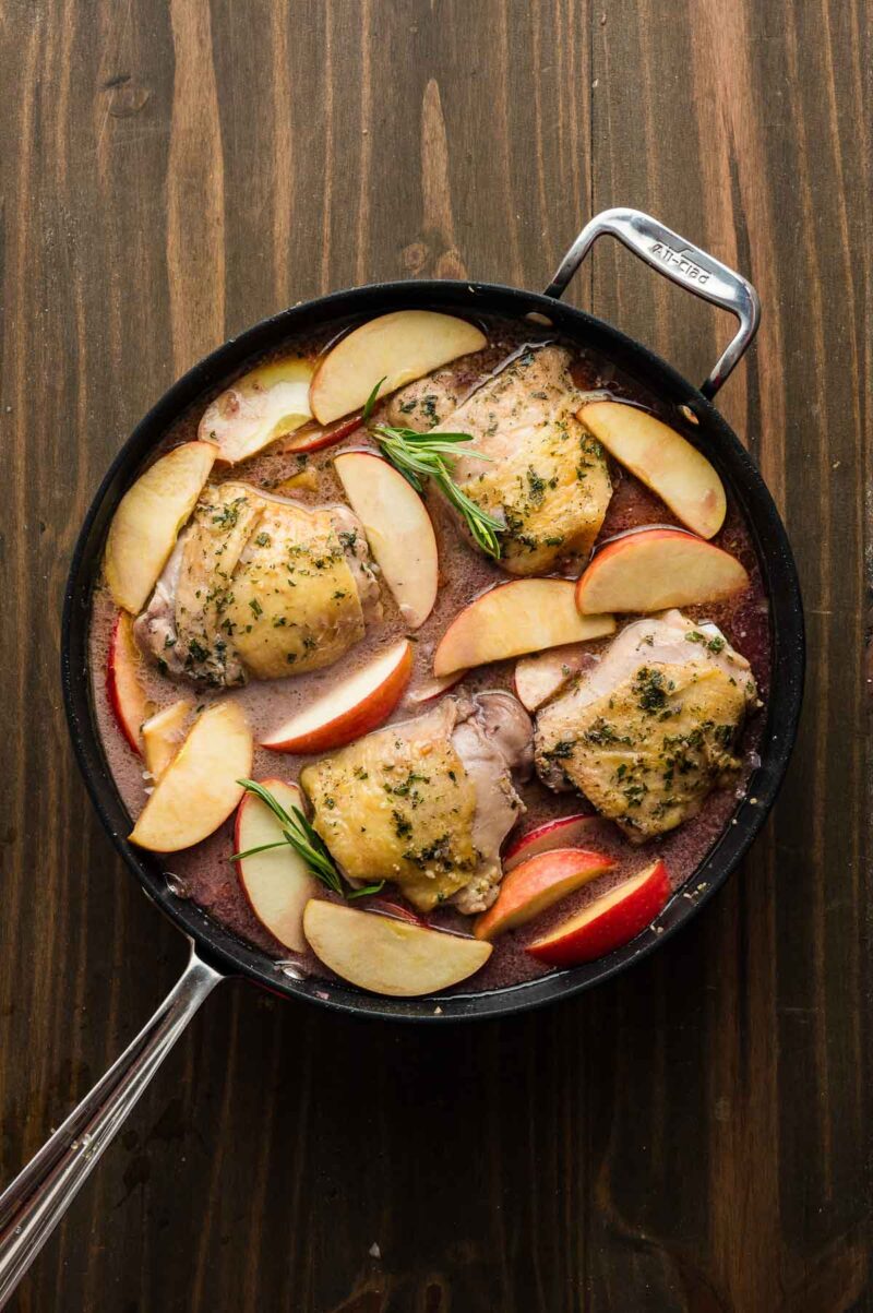 A skillet with cooked chicken thighs, sliced apples, and fresh herbs in a sauce, placed on a wooden surface.