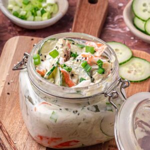 A glass jar filled with creamy cali roll cucumber salad salad containing sliced cucumber, kani, green onions, and sesame seeds, placed on a wooden board with extra vegetables nearby.