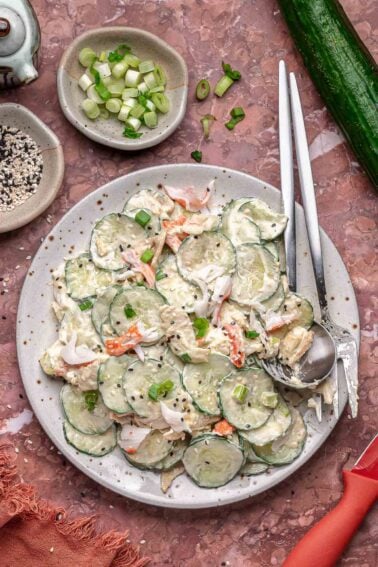 A plate of creamy cali roll cucumber salad with crab, topped with green onions and sesame seeds, surrounded by small bowls of sliced green onions and sesame seeds, with utensils on the side.