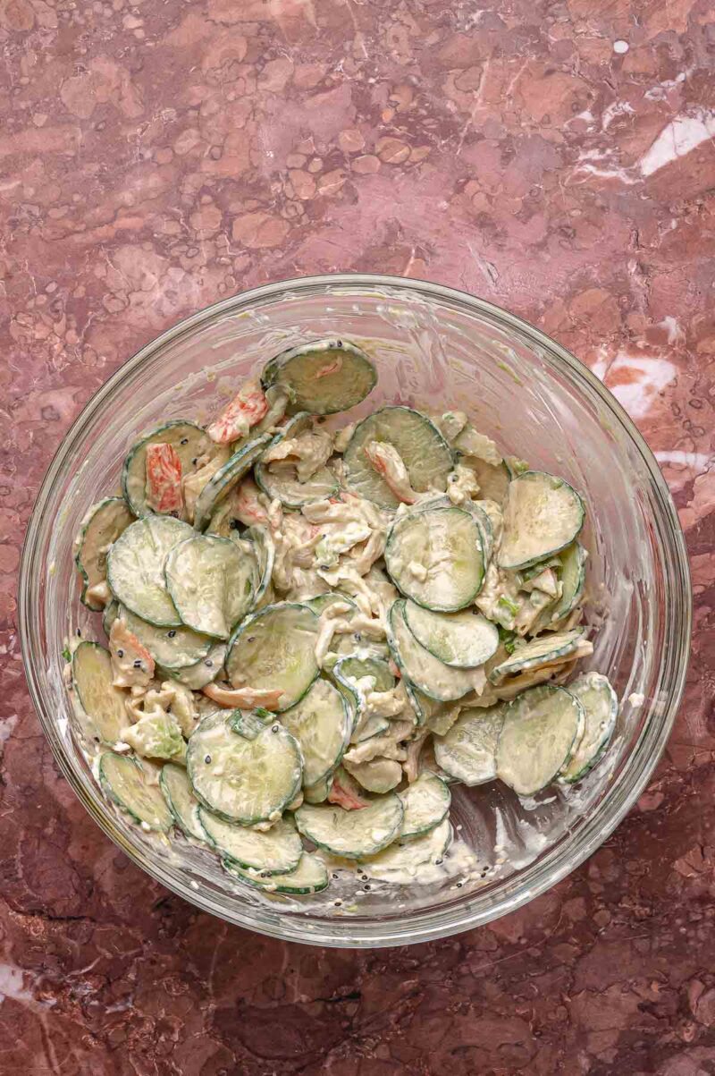 A glass bowl filled with cucumber salad mixed with creamy dressing and sliced vegetables sits on a marbled surface.