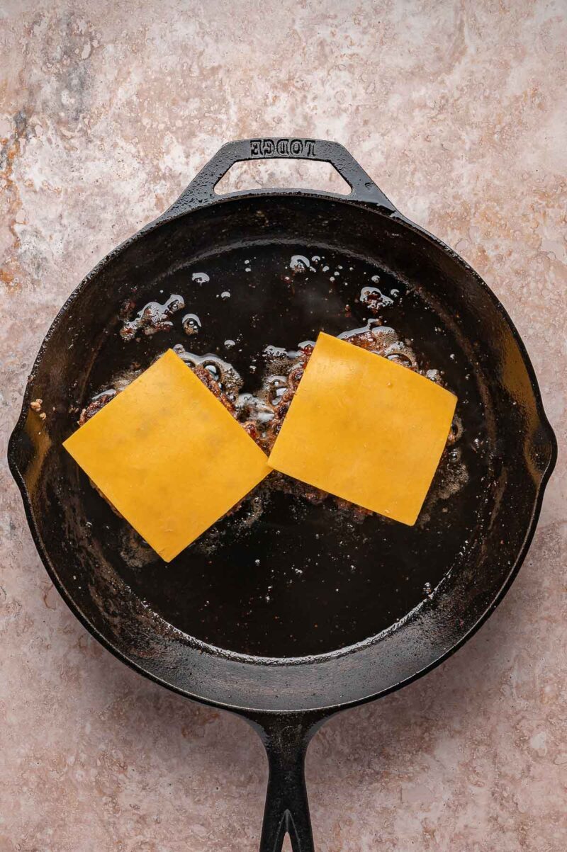 A cast iron skillet with two hamburger patties frying, each topped with a slice of yellow cheese.