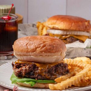 A cheeseburger with lettuce and onion is served on a plate with crinkle-cut fries; another burger and a glass of soda are in the background.