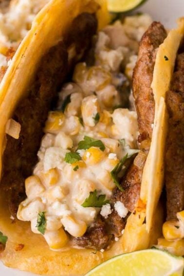 Three smash tacos tacos filled with ground meat, street corn, cheese, and herbs are served on a plate with lime wedges.