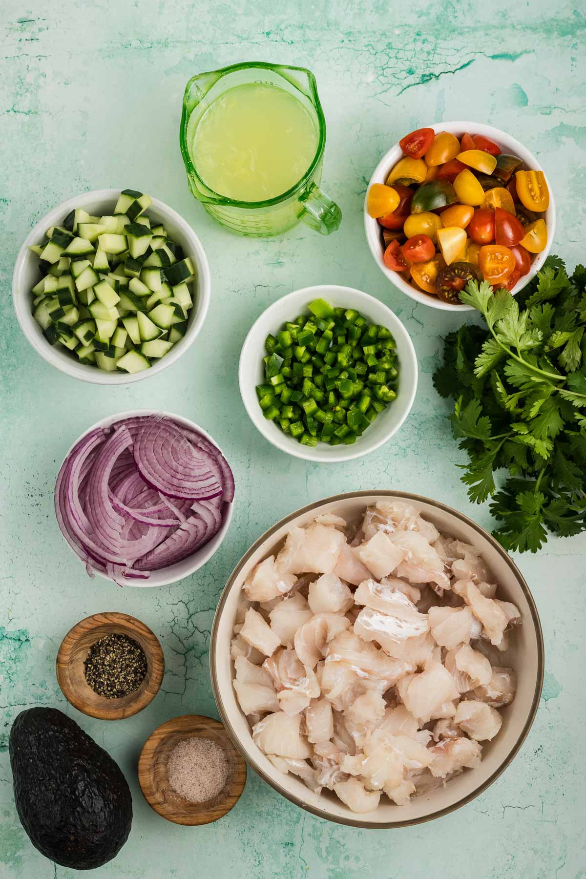 Bowls of diced white fish, cucumber, tomatoes, red onion, jalapeño, cilantro, an avocado, seasonings, and a measuring cup of lime juice are arranged on a light green surface.