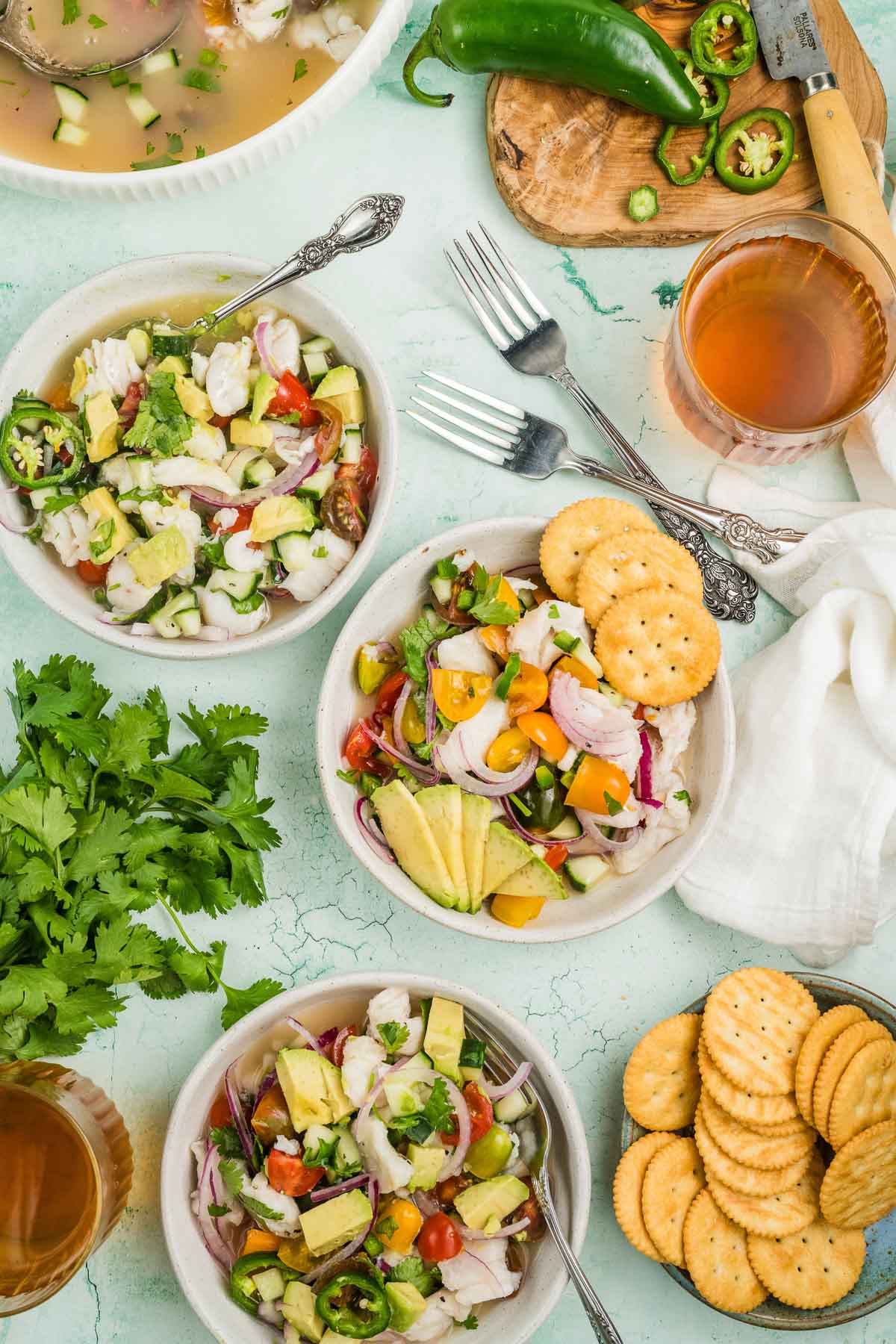 Bowls of ceviche with avocado, tomatoes, and onions are surrounded by round crackers, drinks, sliced jalapeños on a cutting board, and fresh cilantro on a light surface.