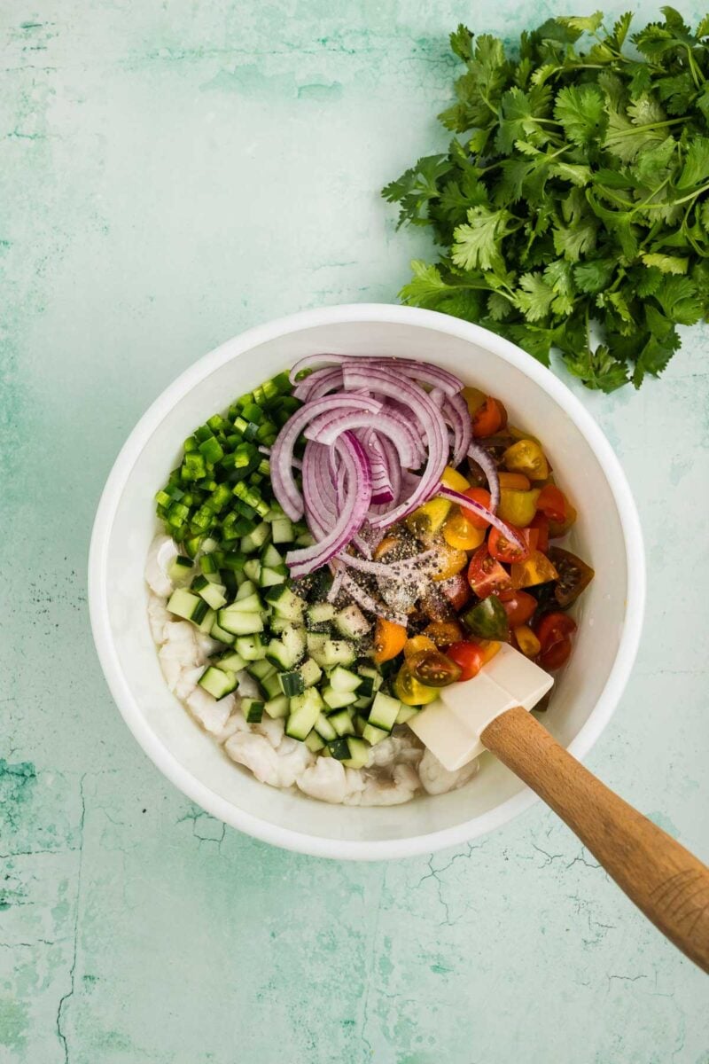 A white bowl filled with chopped shrimp, cucumber, tomatoes, jalapeño, and red onion, with a spatula inside. Fresh cilantro is placed beside the bowl on a green surface.