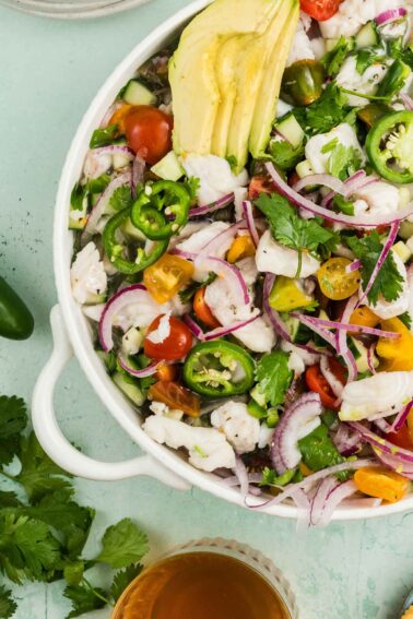 A bowl of seafood ceviche with sliced avocado, red onions, jalapeños, cherry tomatoes, cucumber, and cilantro, served on a light green surface.