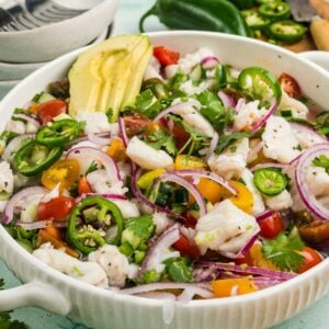 A bowl of ceviche with white fish, red onion, tomato, cilantro, jalapeño slices, and avocado, surrounded by plates and drinks on a light green surface.