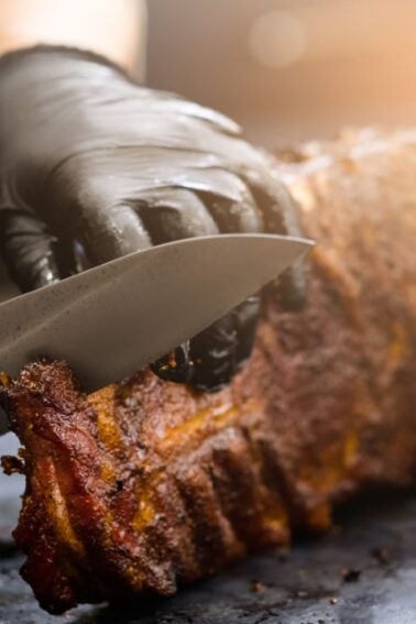 Person wearing black gloves slicing a rack of smoked or grilled ribs on a dark surface.