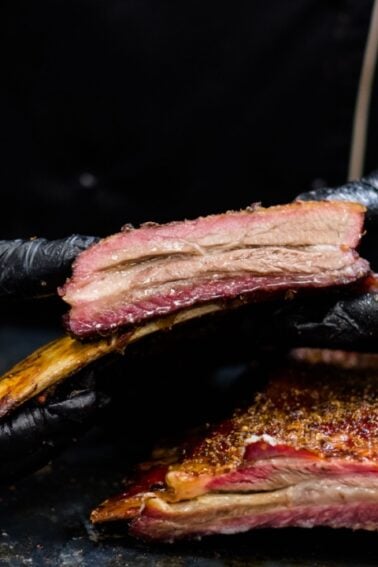 Person wearing black gloves holds and separates smoked beef ribs, showing the meat's texture; a knife and more ribs are visible on the table.