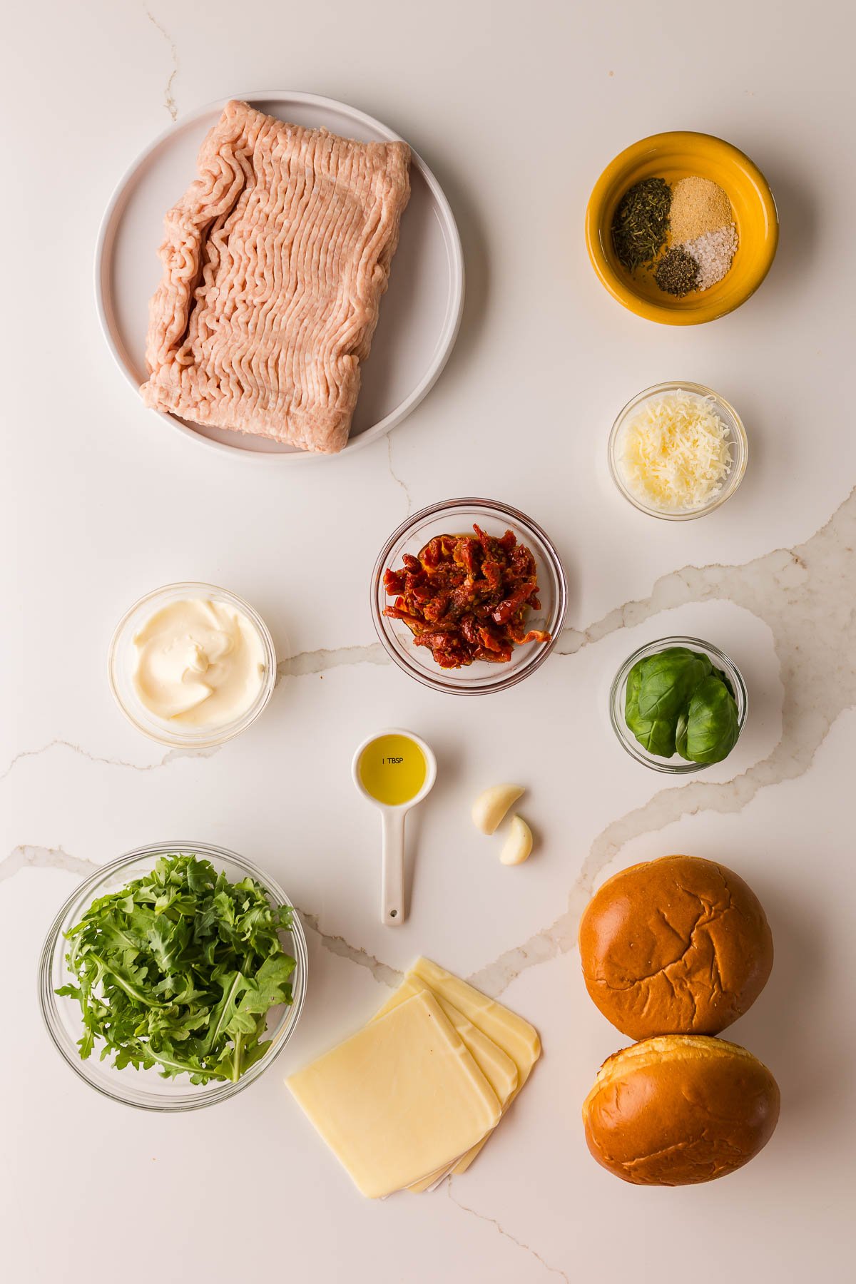 Flat lay of ingredients on a white surface, including ground chicken, seasonings, sun-dried tomatoes, cheese, mayonnaise, basil, arugula, olive oil, garlic, and two burger buns.