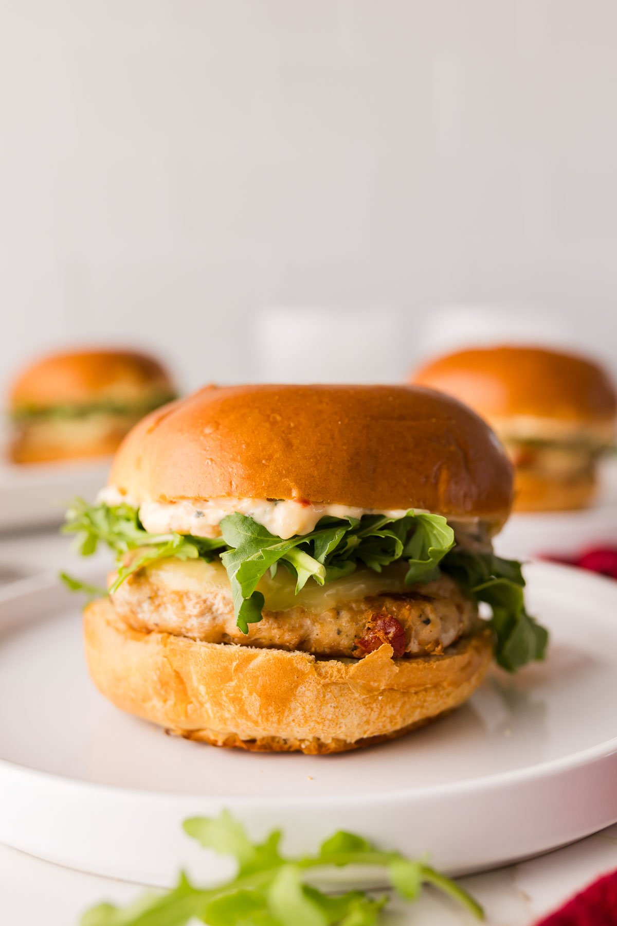 A chicken slider with lettuce and sauce in a brioche bun is served on a white plate, with two similar burgers in the background.