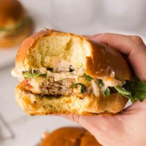 A hand holding a bitten chicken sandwich with lettuce and sauce in a soft bun, with another sandwich blurred in the background.