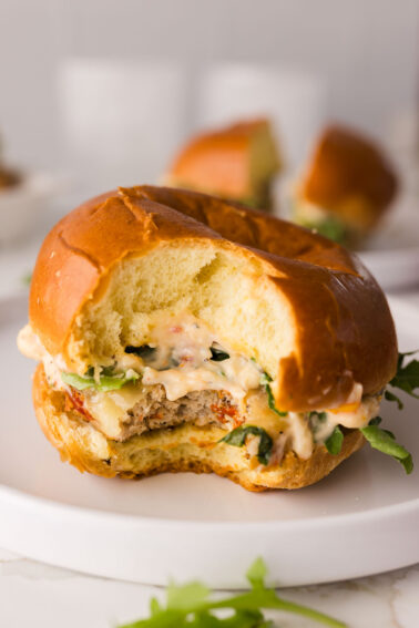 A chicken slider with lettuce and creamy sauce on a brioche bun sits on a white plate, with a bite taken out of it.
