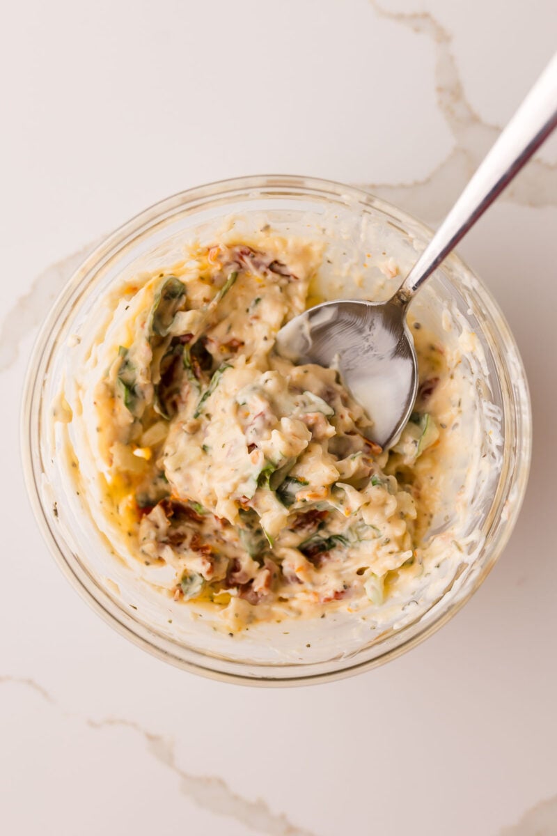 A glass bowl with a mixture of mayonnaise, herbs, and sun-dried tomatoes, with a metal spoon, on a light marble surface.
