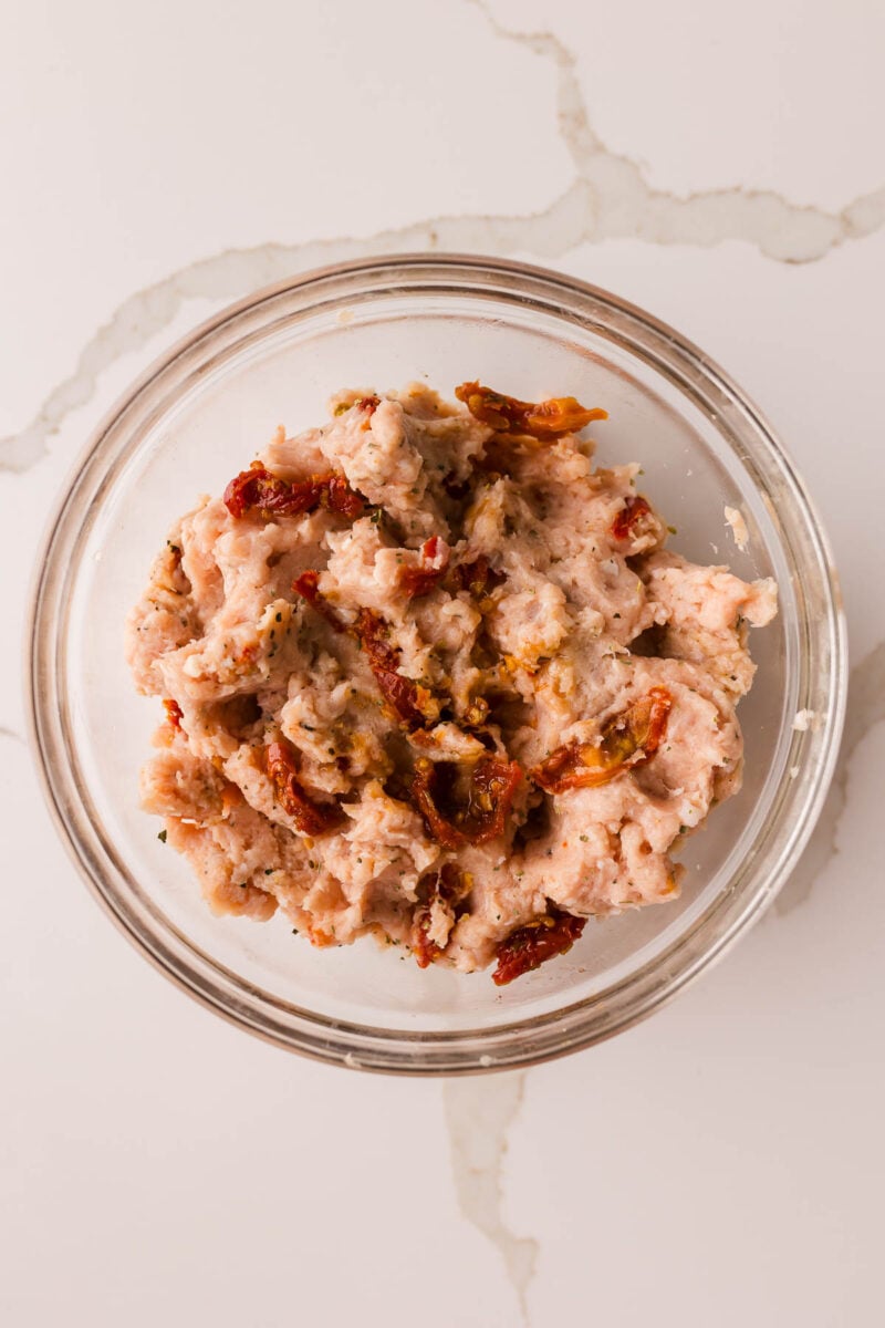 A glass bowl containing ground chicken mixed with pieces of sun-dried tomatoes on a light-colored countertop.