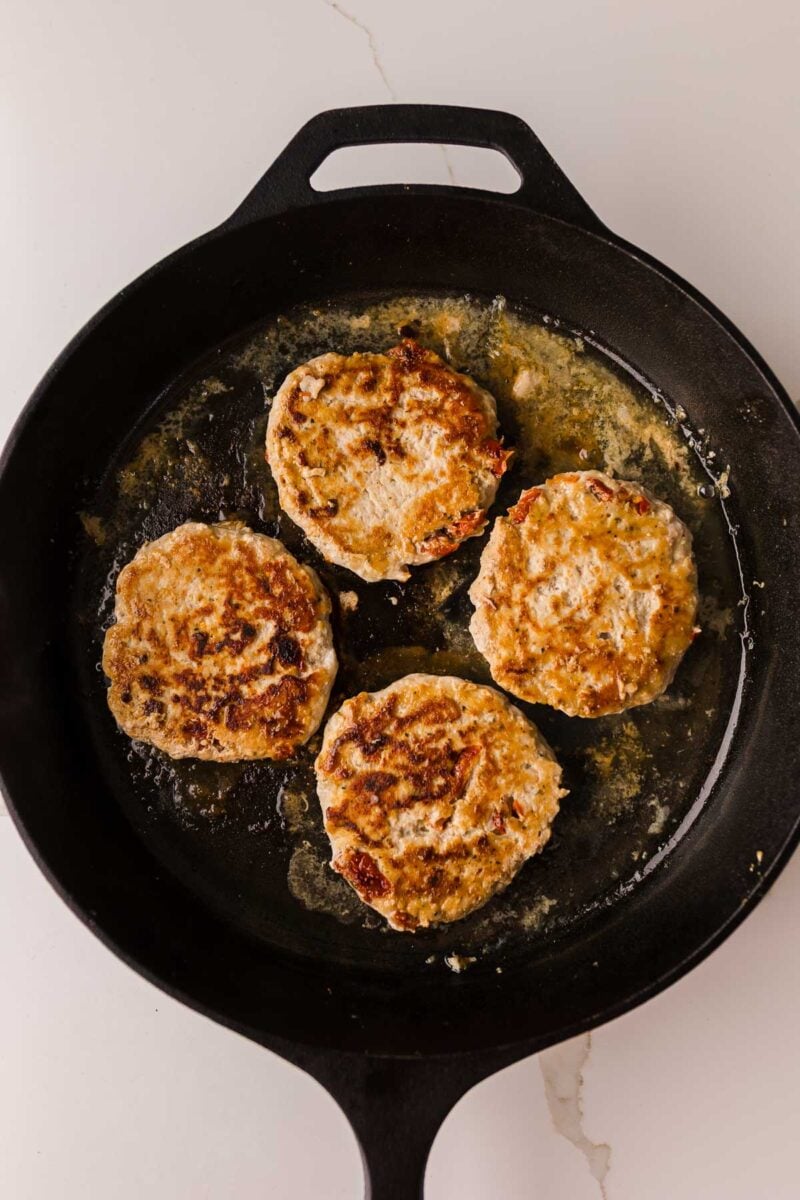Four browned chicken sliders are cooking in a black cast iron skillet, shown from above on a white surface.
