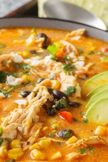 A bowl of chicken tortilla soup garnished with sliced avocado, cilantro, black beans, corn, tomatoes, and shredded chicken, with a spoon and napkin beside it.