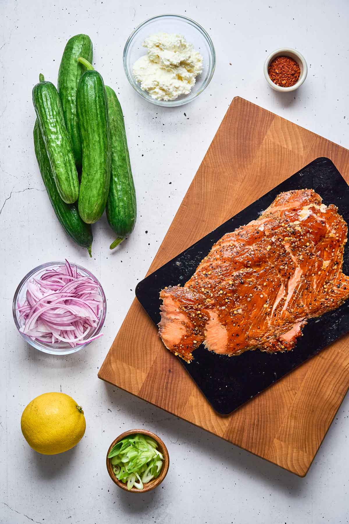 A cutting board with seasoned salmon, surrounded by cucumbers, a bowl of sliced red onions, a lemon, scallions, crumbled cheese, and a small bowl of red spices on a white surface.