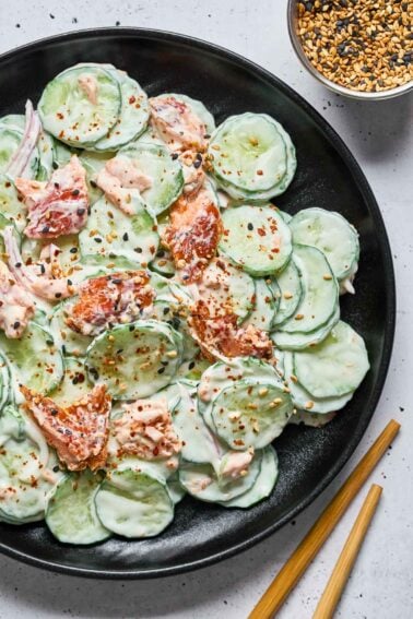 A black plate with creamy Philly roll cucumber salad, pieces of smoked salmon sprinkled with sesame seeds and chili flakes. A bowl of seeds and a pair of chopsticks are beside the plate.