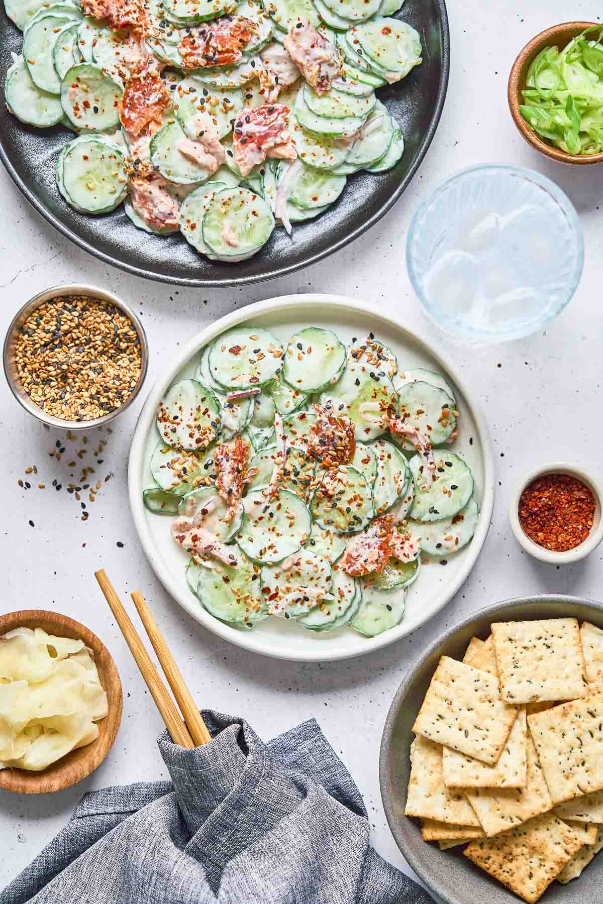 Plate of creamy philly roll cucumber salad topped with red pepper flakes, surrounded by sesame crackers, a bowl of seasoning, lettuce, pickled ginger, and a glass of water on a white surface.