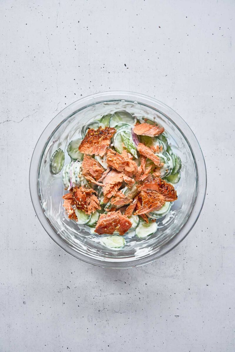A glass bowl containing sliced cucumbers in a creamy dressing, topped with chunks of cooked salmon, on a light gray surface.