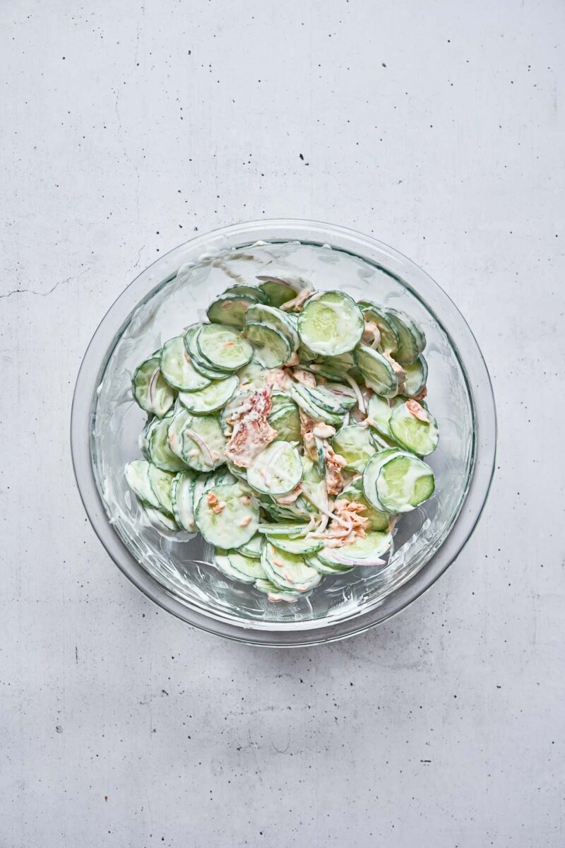 A glass bowl filled with sliced cucumbers and onions mixed in a creamy dressing, placed on a light gray surface.