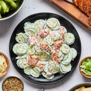 A plate of Philadelphia roll cucumber salad with creamy dressing and seasoning, surrounded by edamame, crackers, flatbread, scallions, pickled ginger, and a bowl of seeds on a white surface.