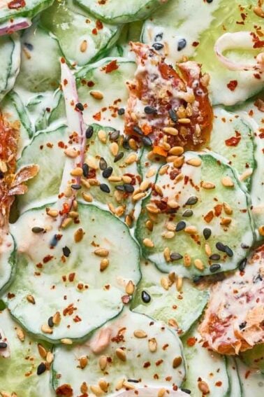 A plate of sliced cucumbers with creamy dressing, tomato pieces, seeds, and red pepper flakes sprinkled on top.
