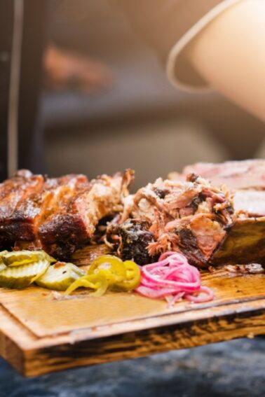 Person wearing black gloves holds a wooden tray with assorted smoked meats and garnishes, including pickles and onions.