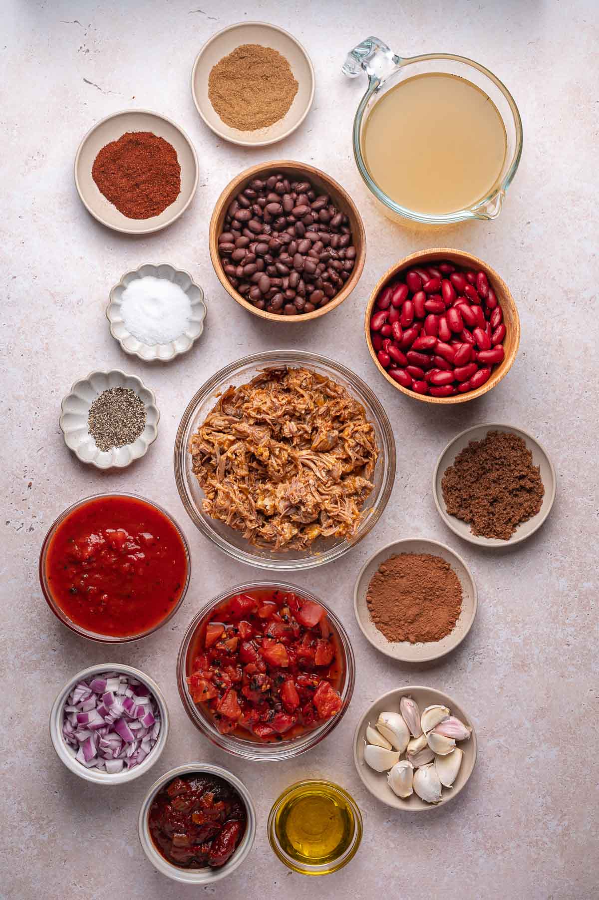 An overhead view of chili ingredients: pulled pork, black beans, kidney beans, spices, broth, tomatoes, salsa, onion, garlic, oil, and chipotle peppers arranged in bowls on a light surface.