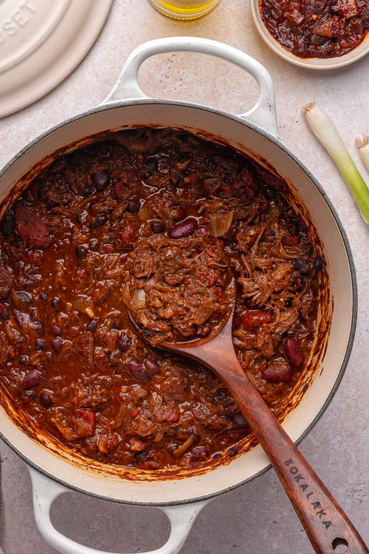 A pot of chili with pulled pork, beans, and tomatoes, with a wooden spoon resting inside. A bowl of chili and some scallions are visible nearby.