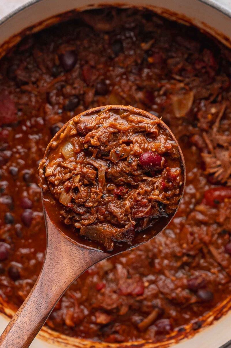 A wooden ladle holds a serving of chunky pulled pork chili with beans, tomatoes, and shredded meat above a pot filled with the same hearty chili.