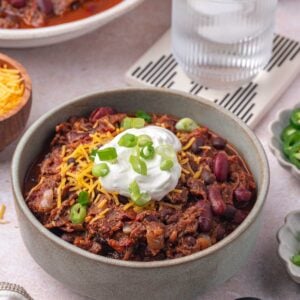 A bowl of chili topped with shredded cheese, sour cream, and sliced green onions, with a spoon, napkin, and a glass of ice water nearby.