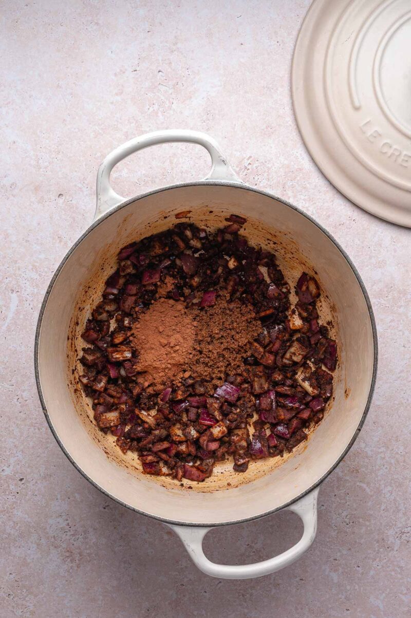 A cream-colored Dutch oven contains sautéed chopped onions and a pile of cocoa powder in the center, ready to become the flavorful base for pulled pork chili; the lid rests nearby on a light countertop.