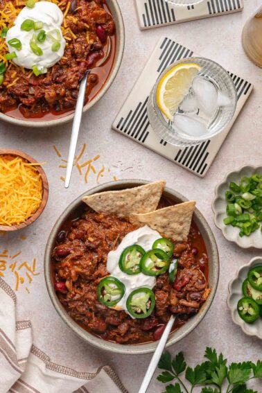 Two bowls of pulled pork chili topped with sour cream, shredded cheese, sliced jalapeños, green onions, and tortilla chips, served with drinks and garnishes on a light table.